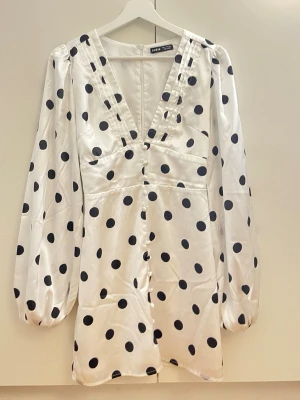 Vit prickig jumpsuit från Zara - Snygg vit jumpsuit från Zara med svarta stora prickar. Djup V-ringning framtill, långa puffärmar och knappar längs framsidan. Tillverkad i ett glansigt, satinliknande material som ger en lyxig känsla. Perfekt för dig som vill sticka ut med en trendig och lekfull look.