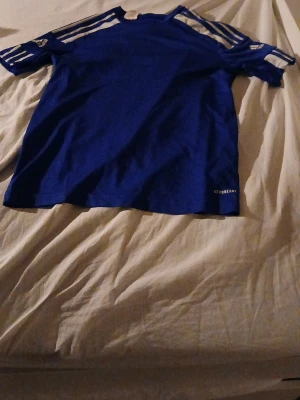 Blå Adidas t-shirt med vita ränder - Säljer en blå Adidas t-shirt med klassiska vita ränder på axlarna och logga på ärmen. T-shirten har korta ärmar och är tillverkad i ett lätt och sportigt material med AEROREADY-teknologi. Perfekt för dig som gillar sportig stil.