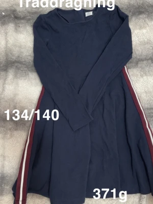 Marinblå långärmad klänning - Mysig marinblå klänning med lång ärm och rund hals. Klänningen har röda och vita ränder längs sidorna som ger en sportig touch. har en ledig passform som passar perfekt till vardags.🌸 Några små trådar hänger ut. Storlek 134/140 