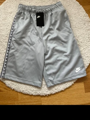 Grå Nike shorts med logga - Snygga grå shorts från Nike med vit logga på benet och svartvit Nike-text längs sidorna. Elastisk midja för skön passform och sportig look. Perfekta för träning eller chill. Tillverkade i mjukt och lätt syntetmaterial.