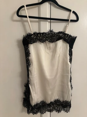 Vit sidenlinnetopp med svart spets - Snygg vit linnetopp i siden med tunna axelband och dekorativ svart spets runt kanterna. Perfekt för dig som vill ha en elegant och trendig look. Toppen har en croppad passform och känns både lyxig och stilren.