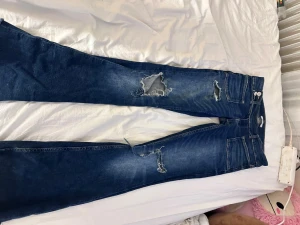 Mörkblå slitna bootcut jeans - Säljer ett par mörkblå bootcut jeans med slitningar och hål på framsidan samt råa, fransiga benslut. Jeansen har klassisk femficksmodell och hög midja. Perfekta för en trendig och avslappnad look.
