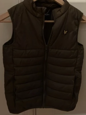 Olivgrön dunväst från Lyle & Scott - Snygg olivgrön dunväst från Lyle & Scott Junior med guldfärgad logga på bröstet. Västen har quiltad design, hög krage och dragkedja framtill. Perfekt för lager-på-lager och ger en sportig vibe till din outfit. Nypris ca 699kr