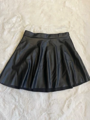 Svart kortkjol i skinnimitation H&M - Säljer en svart kortkjol från H&M i skinnimitation. Kjolen har en lätt utställd form och bred linning som ger en snygg siluett. Perfekt för dig som vill ha en edgy och trendig look.