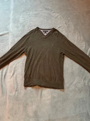 Mörkgrön V-ringad tröja från Tommy Hilfiger - Snygg mörkgrön V-ringad tröja från Tommy Hilfiger. Tröjan har lång ärm, diskret logga på bröstet och är tillverkad i ett tunt, mjukt material som passar perfekt till lager på lager. Klassisk och stilren design som funkar till det mesta.