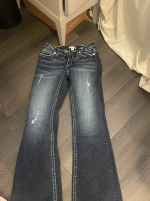 Blå bootcut jeans med broderade fickor - Snygga mörkblå bootcut jeans med slitningar och kontrastsömmar. Baktill finns två fickor med dekorativt broderi i vitt. Jeansen har klassisk femficksdesign och är tillverkade i jeansmaterial med lite stretch för skön passform. Helt nya 
