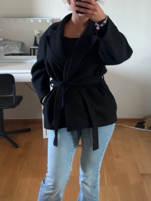 Kappa från missay - Stilren svart finely kappa med knytskärp i midjan och bred krage. Kappan har en rak passform och är tillverkad i ett mjukt tyg som ger en snygg siluett. Perfekt att slänga över en outfit för en clean look.