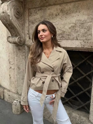 Kort beige trenchcoat med knytskärp - Helt slutsåld trendig kort trenchcoat i beige med dubbelknäppning och knytskärp i midjan. Jackan har klassisk krage, långa ärmar med slejfdetalj och två framfickor. Perfekt för en stilren look under vår och höst. Helt oanvänd med prislapp kvar!