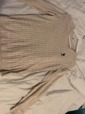 Beige kabelstickad tröja Polo Ralph Lauren - Kabelstickad tröja i beige från US polo assn med klassisk svart logga broderad på bröstet. Tröjan har rund halsringning, långa ärmar och ribbade muddar vid ärmslut och nederkant. Perfekt för lager på lager och enkel att matcha.