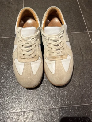 Beige och vita sneakers i mocka från Skolyx - Snygga Skolyx sneakers i beige mocka och vitt läder med klassisk gummisula. Skorna har lågt skaft, vita snören och en retroinspirerad design som passar till många olika stilar. Perfekta för dig som gillar minimalistiska och stilrena sneakers. Orginal pris 1 700kr. Obs låda ingår ej!