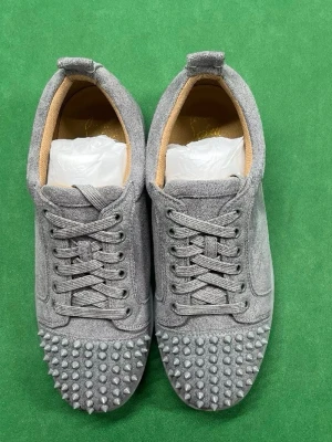 Gråa sneakers med nitar från Louboutin - Unika gråa sneakers från Louboutin i mocka med coola nitar över tån. Skorna har klassisk låg profil, rund tå och matchande grå snörning. Insidan är beige och materialet känns lyxigt och trendigt. Perfekta för dig som vill sticka ut med din stil.