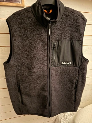 Svart fleeceväst från Timberland - Mysig svart fleeceväst från Timberland med hög krage och dragkedja framtill. Västen har en bröstficka i nylon med dragkedja och Timberland-logga, samt två sidofickor med dragkedja Helt ny säljer den för den var för stor för mig, längden är 71cm så passar M/L.