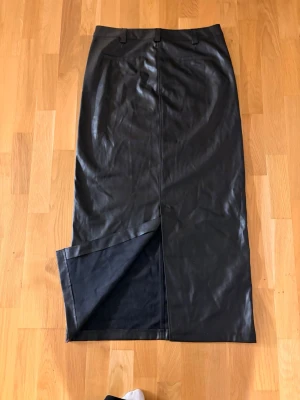 Svart långkjol i skinn från Gina Tricot - Snygg svart långkjol i skinnimitation från Gina Tricot. Kjolen har hög midja, slits baktill och stängs med dragkedja och knapp. Perfekt för dig som vill ha en edgy och trendig look.