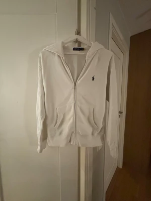 Vit hoodie från Polo Ralph Lauren - Klassisk vit hoodie från Polo Ralph Lauren med dragkedja framtill och två fickor. Ikoniska lilla loggan broderad på bröstet. Tillverkad i mjukt bomullsmaterial och har en skön huva. Perfekt för en avslappnad och stilren look.