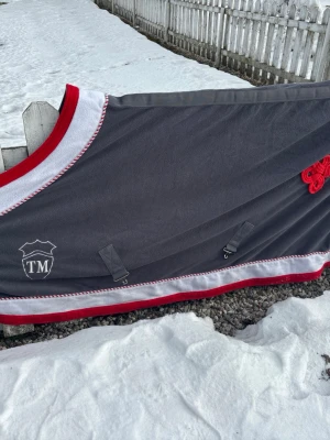 Grå fleecetäcke till häst  - Säljer ett grått fleecetäcke till häst från TM med vita och röda detaljer. Täcket har dubbla spännen framtill, dekorativ kant och broderad logga. 