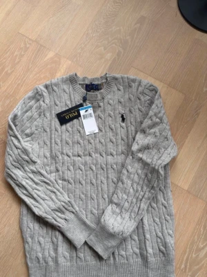 Grå kabelstickad tröja Polo Ralph Lauren - Snygg grå kabelstickad tröja från Polo Ralph Lauren med rund halsringning och klassisk broderad logga på bröstet. Tröjan har ribbade muddar och är tillverkad i mjukt material som passar perfekt till höst och vinter. PRIS KAN DISKUTERAS