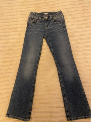 Blå bootcut jeans med broderade fickor - Säljer ett par blå bootcut jeans med snygga slitningar och broderade bakfickor med dekorativa knappar. Jeansen har klassisk femficksmodell, låg midja och är tillverkade i denim med lätt stretch. Perfekta för en avslappnad och trendig look.