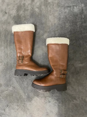 Bruna höga boots med fuskpäls - Snygga bruna stövlar i läderimitation med grov sula och vit fuskpälskant upptill. Stövlarna har två dekorativa spännen i mässing på sidan och en chunky, robust look. Perfekta för kalla dagar och ger en cool vibe till din outfit.