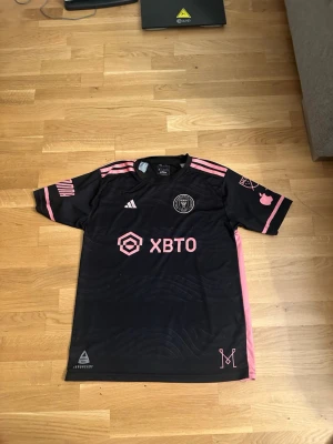 Inter Miami Messi matchtröja svart/rosa - Säljer en svart Inter Miami matchtröja från Adidas med rosa detaljer och tryck. Tröjan har Messi och nummer 10 på ryggen, samt klubbmärke och sponsorlogga framtill. Tillverkad i lätt och ventilerande material, perfekt för fotboll eller träning.