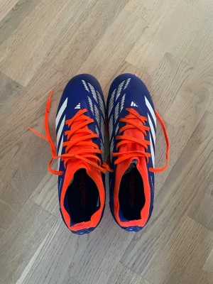 Adidas Predator blå fotbollsskor - Säljer ett par Adidas Predator fotbollsskor i blått med vita detaljer och orange snörning. Skorna har en låg profil och dobbar för spel på gräs. Snygg design med Predator-logga undertill och slitstarkt syntetmaterial för bästa bollkontroll.