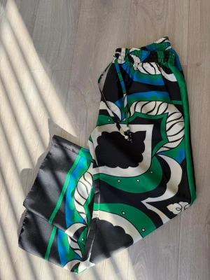 Byxor med grafiskt mönster - Säljer ett par byxor med färgstarkt grafiskt mönster i svart, grönt, blått och vitt. Byxorna har breda ben, resår i midjan och dragsko för skön passform. Materialet är glansigt och känns lyxigt mot huden. Perfekta för dig som vill sticka ut med din stil. Från Zara, helt nya men prislappen sitter ej kvar. 