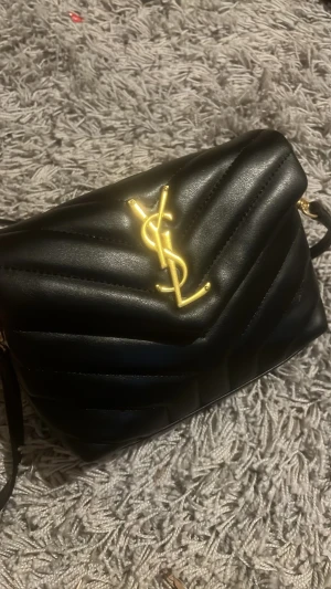 Svart axelväska  - Säljer en svart axelväska med quiltad design och ikoniskt YSL-emblem i guld på framsidan. Väskan har en stilren form och justerbar axelrem. Tillverkad i slätt läder som ger en lyxig känsla.