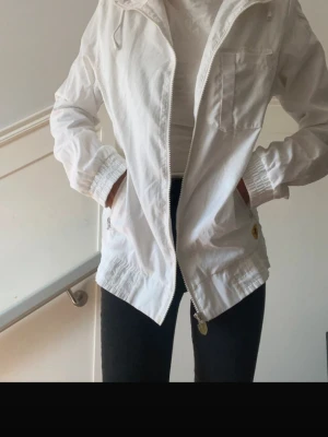 Vit vindjacka från Ferrari - Snygg vit vindjacka från Ferrari med dragkedja, bröstficka och två sidofickor. Jackan har elastiska muddar vid ärmslut och nederkant samt en gul Ferrari-logga vid fickan. Tillverkad i lätt material, perfekt för blåsiga dagar.