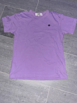 Lila t-shirt från Comme des Garçons - Säljer en lila t-shirt från Comme des Garçons Play med det klassiska lilla hjärtemärket på bröstet. T-shirten har korta ärmar och en avslappnad passform. Perfekt för dig som gillar stilrena plagg med en twist.
