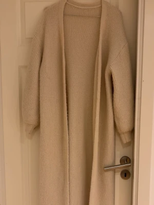 Lång beige stickad kofta - Mysig lång kofta i beige stickat material med öppen front och långa ärmar. Perfekt för lager-på-lager och enkel att matcha med olika outfits. Koftan har en avslappnad passform och ribbstickade muddar vid ärmsluten.