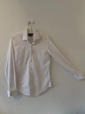 Vit slim fit skjorta från Dressmann - Klassisk vit skjorta från Dressmann i slim fit-modell. Skjortan har långa ärmar, knappar framtill och en snygg krage. Perfekt för en clean och stilren look.