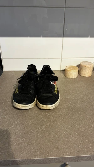 Svarta Dsquared2 sneakers med tryck - Snygga svarta sneakers från Dsquared2 med vita sulor och svart snörning. Skorna har gul text på sidan och ett rött lönnlöv under sulan. Tillverkade i läder med klassisk låg profil och coola detaljer som sticker ut. Finns en slitning på ena skon vid hälen, inget som syns när man har på sig dom men om man vill fixa det så kostar de typ 200-300 hos en skomakare :D