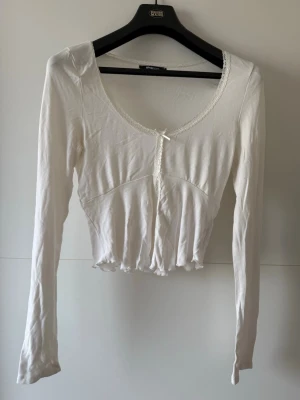 Vit croppad topp med spetsdetaljer - Söt vit croppad topp från Shein med långa ärmar, spetskant runt ringningen och en liten rosett framtill. Toppen har en figurnära passform och vågig kant nertill. Perfekt för att skapa en romantisk och trendig look.