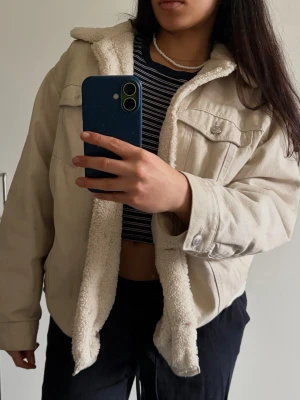 Beige teddyfodrad cardiganjacka - Mysig beige cardiganjacka med vitt teddyfoder på insidan. Jackan har krage, knappstängning och två bröstfickor med lock. Perfekt för lager på lager och ger en avslappnad look. Materialet är mjukt och värmande, perfekt för kyligare dagar.