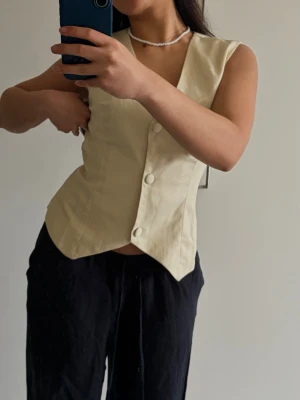 Beige väst med knyt och knappar - Stilren beige väst med v-ringning och stora knappar framtill. Västen har ett snyggt knytband i ryggen som ger en feminin siluett. Tillverkad i ett lätt material, perfekt för lager på lager. Passar till både jeans och kjol.