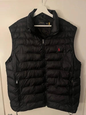 Svart dunväst från Polo Ralph Lauren - Snygg svart dunväst från Polo Ralph Lauren med klassisk quiltad design och röd broderad logga på bröstet. Västen har hög krage, dragkedja framtill och två fickor med dragkedja. Perfekt för lager-på-lager och pris kan diskuteras vid snabb affär