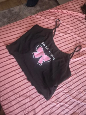 Svart croppad linnetopp med rosett - Söt svart croppad linnetopp med tunna axelband och ett tryck av en rosa rosett samt texten 'good night' framtill. Perfekt för en avslappnad och trendig look.