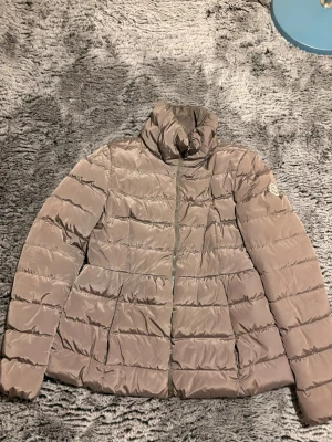 Grå. pufferjacka från Moncler - Säljer en grå pufferjacka från Moncler med hög krage och dragkedja framtill. Jackan har två sidofickor och det klassiska Moncler-märket på ärmen. Tillverkad i glansigt material som ger en modern look och perfekt för kalla vinterdagar.