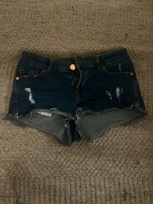 Mörkblå jeansshorts med slitningar - Säljer ett par mörkblå jeansshorts med råa kanter och slitna detaljer framtill. Shortsen har klassisk femficksdesign, knapp och dragkedja i gylfen samt bälteshällor. Perfekta för en avslappnad och trendig look.