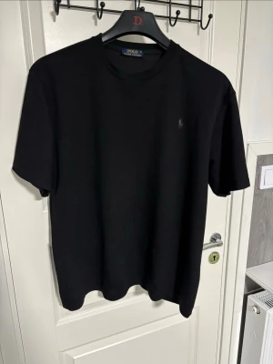 Svart t-shirt från Polo Ralph Lauren - Klassisk svart t-shirt från Polo Ralph Lauren med diskret broderad logga på bröstet. T-shirten har rund halsringning och korta ärmar, perfekt för en stilren och enkel look.