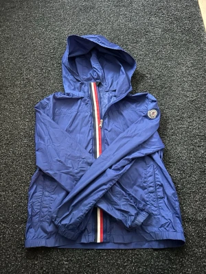 Moncler Windbreaker - Snygg blå vindjacka från Moncler med huva och dragkedja framtill. Jackan har Moncler-logga på ärmen och en randig detalj längs dragkedjan i rött, vitt och blått. Tillverkad i lätt och tåligt material, perfekt för blåsiga dagar. Passar runt 160 cm. Hojta till vid funderingar och liknande😊hör av er innan köp tack 