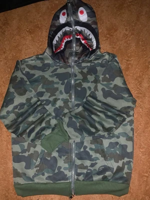 BAPE camo hoodie med hajmotiv - Säljer en grön camo hoodie från BAPE med dragkedja och ikoniskt hajmotiv på huvan. Hoodien har ribbade muddar och fickor framtill. Perfekt för dig som vill sticka ut med streetwear-stil. Materialet är mjukt bomullstyg.