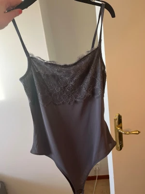 Mörkgrå spetsbody med tunna axelband - Snygg mörkgrå body med tunna axelband och vacker spets upptill. Perfekt för att skapa en stilren och feminin look. Materialet är mjukt och följsamt, och spetsen ger en extra touch av elegans.