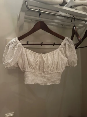 Vit broderad croppad blus - Söt vit croppad blus med puffärmar och broderade detaljer. Toppen har smockad midja, offshoulder-modell och en liten knytning framtill. Perfekt för en trendig och somrig look. Köpare står för frakt. Skriv vid frågor eller intresse💗