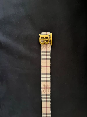 Beige rutigt Burberry-bälte - Snyggt bälte från Burberry med klassiskt beige, svart och rött rutmönster samt guldfärgat spänne med logga. Bältet har detaljer i skinn och den ikoniska riddaren tryckt på bandet. Perfekt accessoar för att lyfta din outfit.