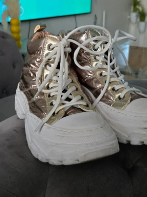 Chunky metallic guld sneakers - Snygga sneakers med metallic guldglans och vita tjocka sulor. Skorna har högt skaft, vita snören och quiltad textil på ovandelen. Perfekta för dig som vill sticka ut med en trendig och unik look.