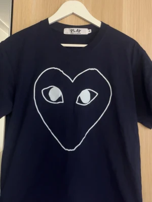 Mörkblå  t-shirt Comme des Garçons Play - mörkblå t-shirt från Comme des Garçons Play med det ikoniska hjärtmotivet med ögon på bröstet. Klassisk passform och rund halsringning. Perfekt för dig som gillar streetwear och vill sticka ut med en stilren design.