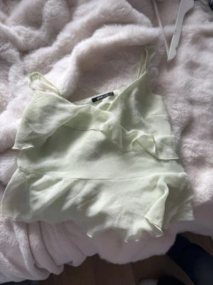Ljusgrön linnetopp från Gina Tricot - Söt ljusgrön linnetopp från Gina Tricot med tunna axelband och volangdetaljer framtill. Toppen har en luftig och lätt struktur, perfekt för varma dagar. Passar dig som gillar en feminin och trendig stil.