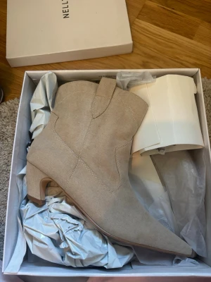 Beige cowboy boots från Nelly - Stilrena beige cowboy boots från Nelly med spetsig tå och låg klack. Tillverkade i mockaliknande material med klassiska sömmar och dragflikar upptill. Aldrig använda och kvar i orginalförpackning 