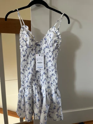Vit blommig kortklänning XS - Supersöt vit kortklänning från Zara med blått blommönster, smockad överdel och tunna axelband. Klänningen har volangkant nedtill och en feminin, figurnära passform. Perfekt för varma dagar och somriga vibes!