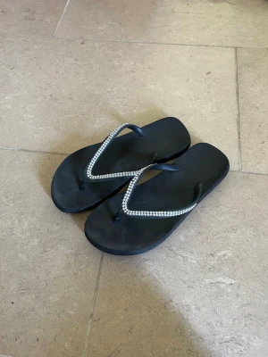 Svarta flip-flops med strassdetaljer - Snygga svarta flip-flops med tunna remmar dekorerade med glittrande strass. Perfekta för sommaren och strandhäng. Klassisk form med platt sula och enkel design som passar till många outfits.